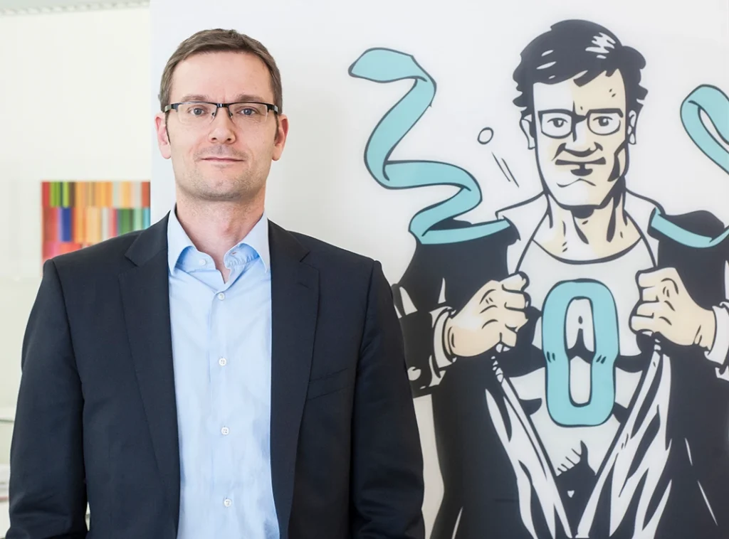Der Vertrauensanwalt Herr Dr. Carsten Thiel von Herff steht neben einer Zeichnung von ihm als Superheld, auf dessen Brust ein O steht.